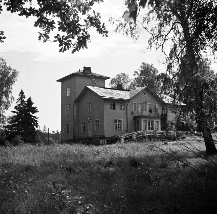 Svartvitt fotografi av Nordsjö gård. I förgrunden lövträd, i mitten ett putsat tvåvåningars trähus. I bakdelen av huset till vänster ett torn på tre våningar. 