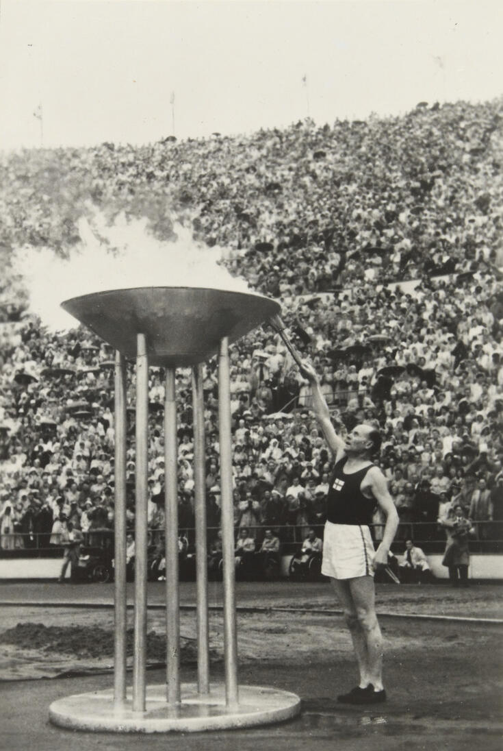 Paavo Nurmi lights the Olympic flame at the Helsinki Olympic stadium. The flame is on a large cauldron, which rests high on five metal poles.