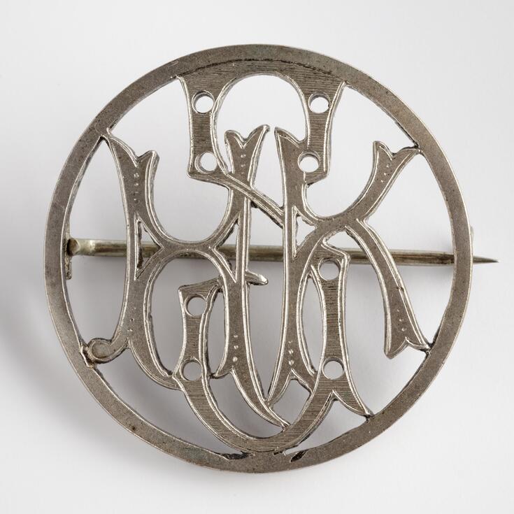 Hopeanvärinen rintaneula, jossa kaunokirjaimilla lomittain kirjaimet HSK