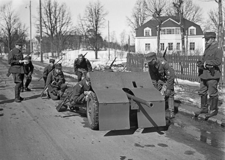 Sotilaita tiellä suuren aseen kanssa. Taustalla huvila ja paljaita lehtipuita.