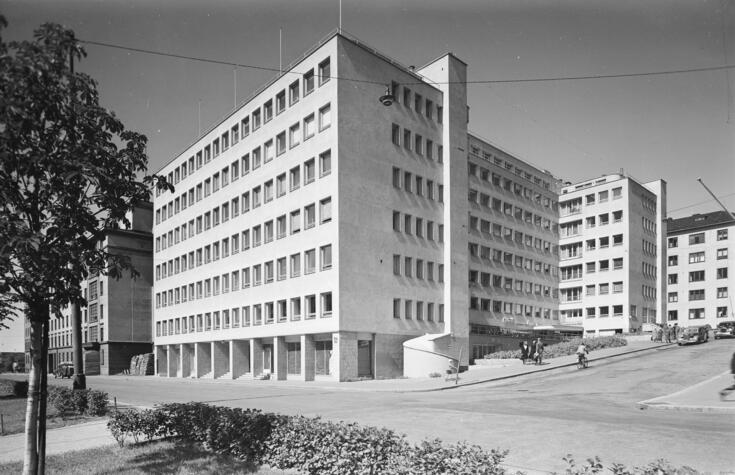 Vaalea kerrostalorakennus. Edessä pohjakerroksessa nelikulmaiset pylväät. Toisella sivulla terassi.