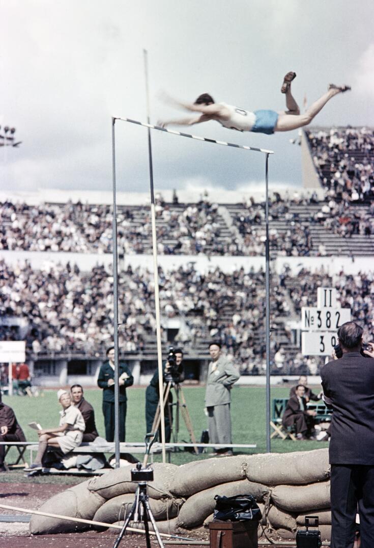 Seiväshyppykilpailu Olympiastadionilla. Urheilija on hyppynsä lakikorkeudessa, juuri riman ylittäneenä.