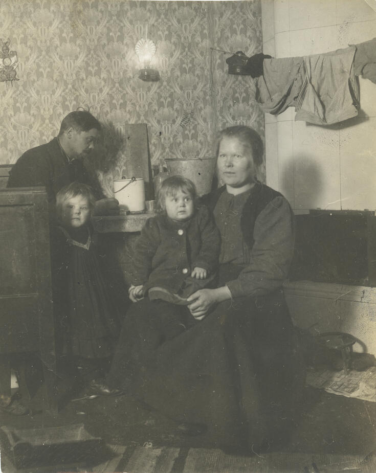Työläisperhe (mies, nainen ja kaksi pientä tyttöä) pienessä asunnossaan n. vuoden 1910 tienoilla. Kakluunin eteen on ripustettu pyykkinaru, jolla on vaatteita kuivumassa.