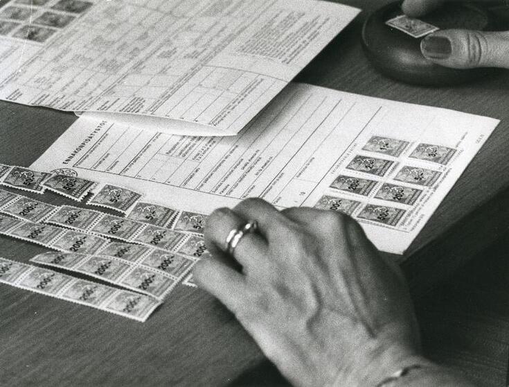 På skrivbordet finns skatteblanketter, skattemärken och en persons händer.