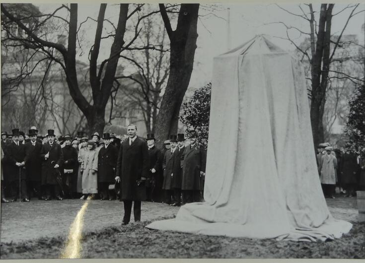 Till vänster står en man i mörk kostym, till höger står ett monument som är övertäckt med vitt tyg.