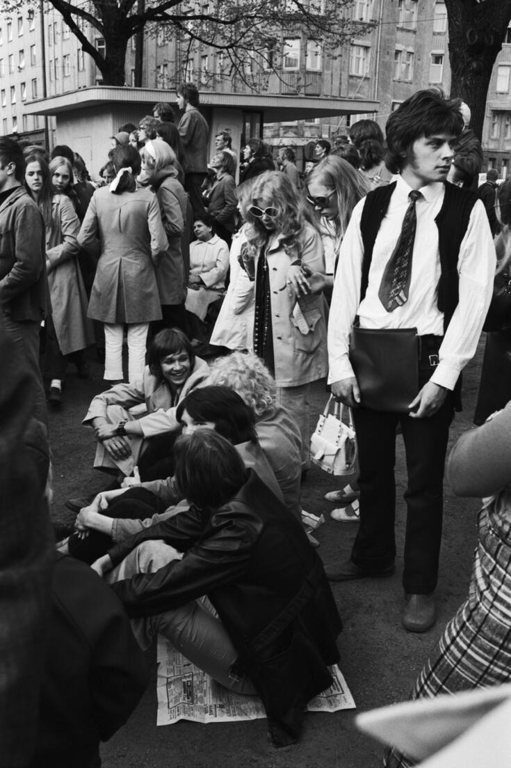 Paljon nuoria kokoontuneena puistoon. Suurin osa seisoo, mutta jotkut istuvat maassa sanomalehtien päällä. Taustalla näkyy lippakioski, pari puunrunkoa ja puistoa ympäröiviä kerrostaloja.