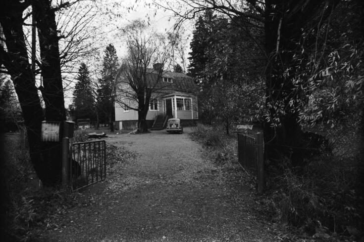 Etualalla on avoin portti, jonka takana aukeaa puiden reunustama syksyinen pihapiiri. Pihassa on puolitoistakerroksinen mansardikattoinen puutalo, jonka edessä on Fiat 600 -henkilöauto.