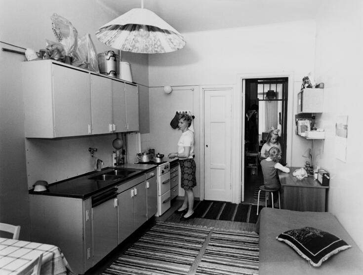 Vasemmalla seinustalla on keittiön kiinteät kalusteet. Nainen seisoo lieden ääressä. Oikealla on kaksi lasta. Taustalla ovi muualle asuntoon.
