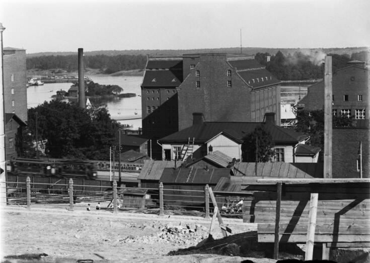 Näkymä Pengerkatu 15:n kohdalta Hämeentie 13:n ja 15:n suuntaan ja Sörnäisten rantaan sekä saarille (vas. Kana, oik. Sompasaari). Oikealla taustalla tiilirakennusten välissä kulkee Lintulahdenkatu, vasemmalla edessä näkyy kerrostalon Pengerkatu 17:n (Sakarinkatu 3) reunaa. Vasemmalla aidan toisella puolella työmaa Hämeentie 38:n tontilla.