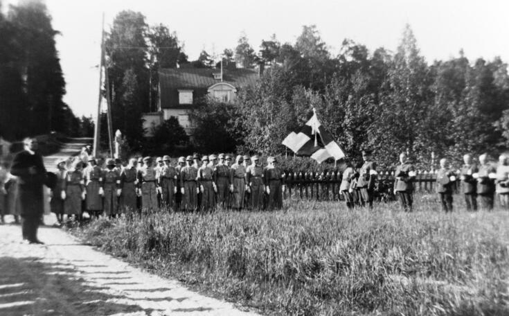 I ett lantligt sommarlandskap står tiotals kvinnor i lottadräkt och skyddskårister, varav en håller en bärbar flaggstång med Finlands flagga.