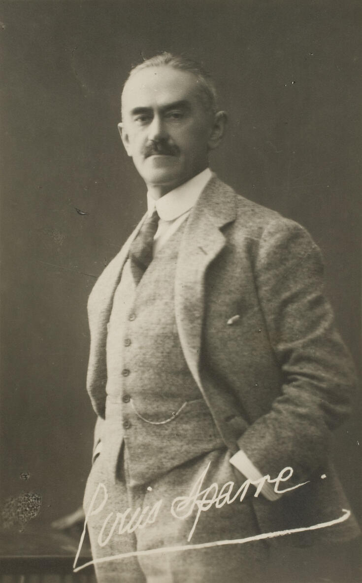 Vaaleaan villakangaspukuun pukeutunut mies seisoo pöydän vieressä vasen käsi taskussaan. Kuvan alareunassa on hänen nimikirjoituksensa. 