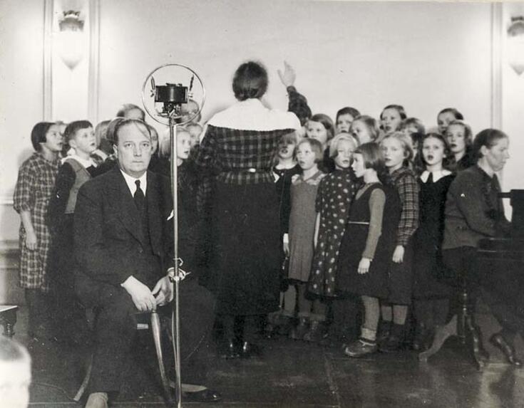 Etualalla istuu pukuun pukeutunut mies. Hänen takanaan on lapsikuoro, jota johtaa nainen selin kameraan. Oikealla istuu nainen pianoa soittamassa.