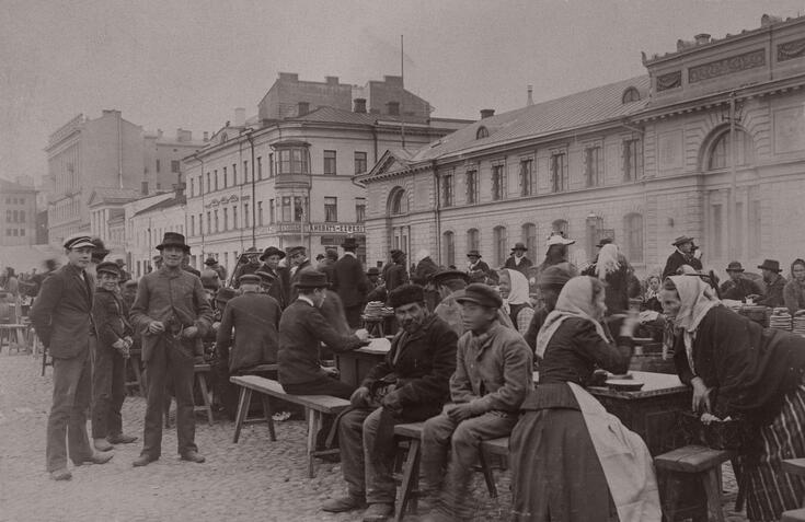 Rahvaan pukuihin pukeutuneita ihmisiä kokoontuneena penkeille pöytien ääreen. Osa ihmisisitä seisoo. Taustalla näkyy kaksi- ja kolmekerroksisia rakennuksia, joita ei enää ole.