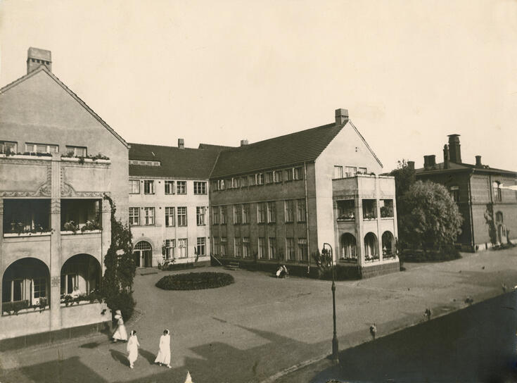 Kolmikerroksisten kivirakennusten ympäröimä sisäpiha, jossa on istutuksia ja penkkejä. Kolme valkopukuista hoitajaa kävelee pihalla. 