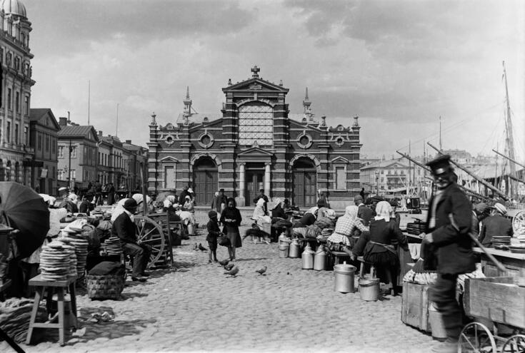 Ihmisiä on kokoontunut kauppahallin Ullanlinnan puoleisen päädyn ulkopuolelle, suurin osa istuu pöydän ääressä. Maassa on maitotonkkia ja muita peltisiä kuljetusastioita sekä kärryjä. Oikealla näkyy purjealuksia.