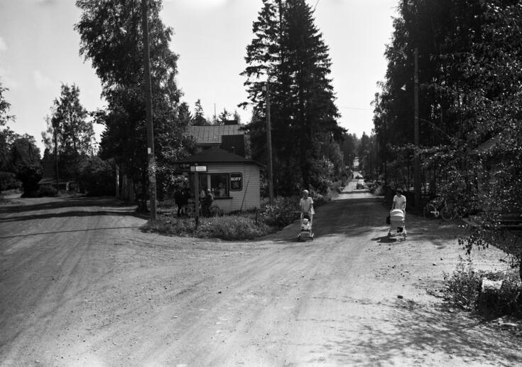 Etualalla on hiekkatie, joka haarautuu vasemmalle ja oikealle. Keskellä on puinen kioskirakennus, jonka edustalla on viisi ihmistä ja kaksi polkupyörää. Teiden reunoilla on lehtipuita. Oikeanpuoleisella tiellä on nainen ja mies, jotka kumpikin työntävät lastenvaunuja. Heidän takanaan näkyy kuutisen autoa, joista osa on pysäköity.