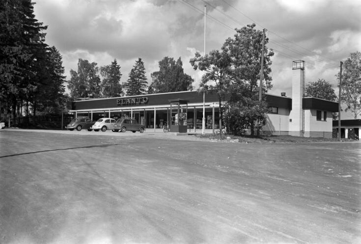 Matala laatikkomainen valkoinen myymälärakennus, jonka yläosassa lukee ELANTO. Rakennuksen edessä on puhelinkoppi ja kaksi pysäköityä Kupla-Volkkaria sekä muu pikkuauto, jonka katolla on tavaroidenkuljetusteline.