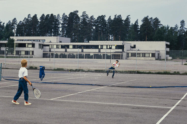 Kaksi nuorta pelaa tennistä. Kenttä on Jakomäen liikuntapuistossa, Suurmetsäntie 5. Taustalla näkyy Jakomäen peruskoulun rakennus, Suurmetsäntie 3. Valokuva on otettu luoteeseen.