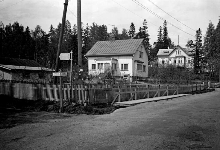 I förgrunden löper en grusväg. I mitten finns en egnahemshustomt omgiven av ett staket. På tomten står ett frontmannahus och en låg gårdsbyggnad. I bakgrunden syns en äldre trävilla. På bilden syns en vägskylt där det står Eskosvägen och Vallmovägen.