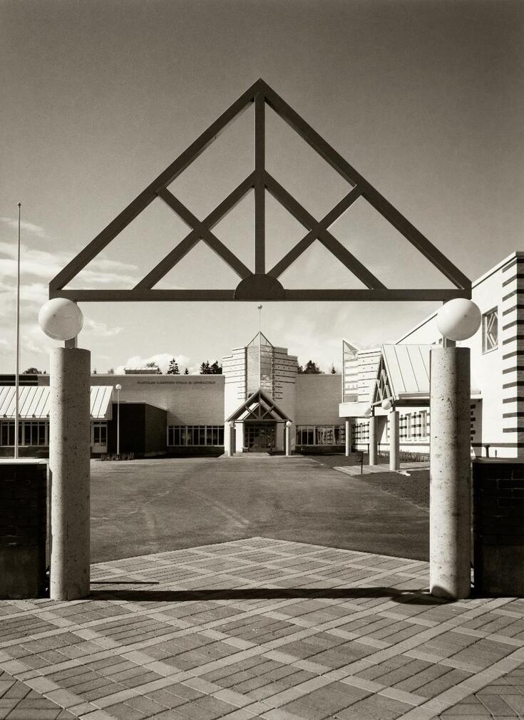Pihaportti, jonka pyöreiden betonipylväiden päällä lepää kolmio. Portin takana on koulun piha ja matalat vaaleat koulurakennukset, joiden sisäänkäyntien yläpuolella toistuu portin kolmioaihe.