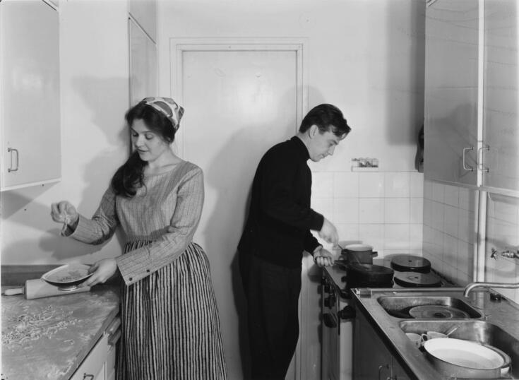 Vasemmalla on nainen keittiön työtason ääressä, oikealla mies lieden äärellä.