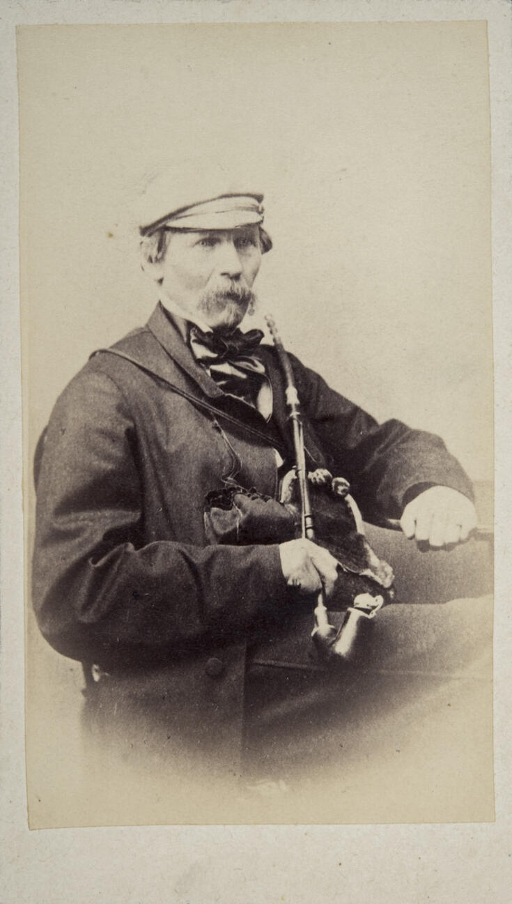 Ulkovaatteisiin pukeutunut mies istuu tuolilla iso, pitkävartinen piippu sylissään