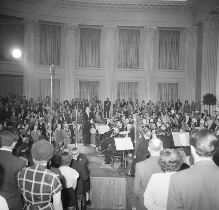 I förgrunden ser man en konsertpublik med ryggen mot kameran, och i bakgrunden en orkester med kapellmästare i universitetets festsal.