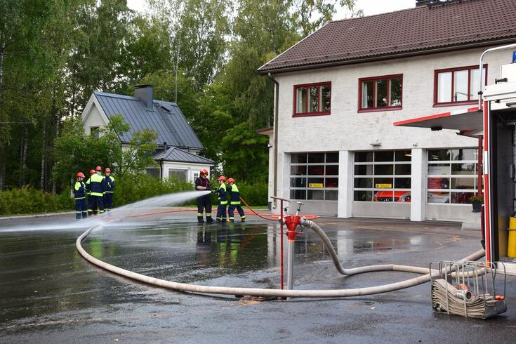 Palokunnan nuoret harjoittelevat toimitilojen pihalla. Sammutusletku on vedetty paloautosta ulos ja vanhempi palomies johtaa toimintaa. Taustalla näkyvissä kolme autotallia, joissa ajoneuvokalustoa. Puistolan Vapaaehtoinen Palokunta r.y. (Asema 53) toimii Helsingin kaupungin pelastuslaitoksen alaisena sopimuspalokuntana.