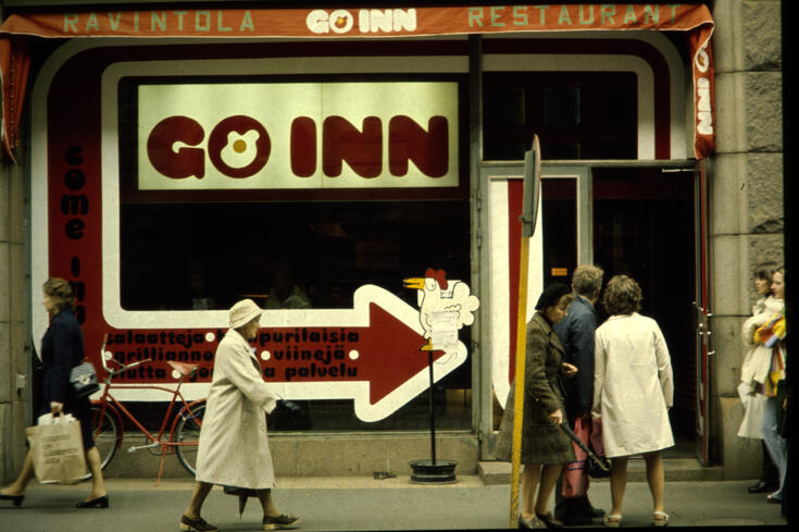 Restaurangens fönster har tejpats med texten GO INN i rött mot vit bakgrund och en stor röd, vitkantad pil som pekar mot restaurangens dörr. Människor promenerar på trottoaren, några syns i dörren till stället. Bredvid ingången står en reklamskylt i form av en höna. På den röda pilen har man skrivit med svart GO INN RAVINTOLA RESTAURANT. Come inn, salaatteja, hampurilaisia, grilliannoksia, viinejä, olutta.