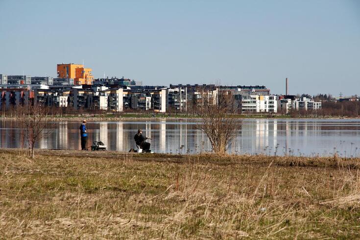 Kaksi kalastajaa rannassa tyynellä lahdella. Taustalla Arabianrannan uusia asuintaloja.
