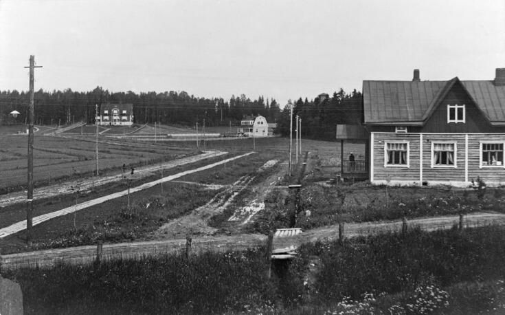 Peltoja, kapeita hiekkateitä ja kolme puurakennusta, joista yhdessä on kauppa. Sen kuistilla seisoo ihminen. 