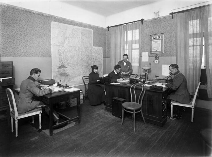 Neljä miestä ja nainen työnteossa. Nainen kirjoittaa kirjoituskoneella. Yksi miehistä puhuu puhelimessa, kaksi keskustelee keskenään. Kuvassa keskellä seisova henkilö on O.A. Östman. Seinällä on suuri kartta.