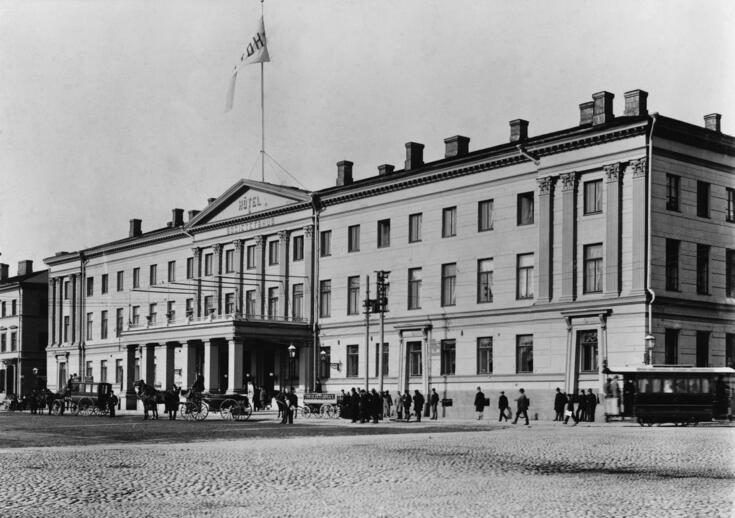 Hotelli Seurahuone kuvattuna Kauppatorilta. Rakennuksen edustalla on jalankulkijoita ja hevosvaunuja. Raitiovaunu on kääntymässä Katariinankadulta Pohjoisesplanadille.