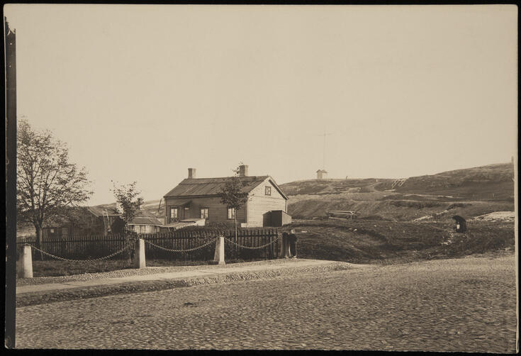 I förgrunden är en kullerstensgata. I mitten är ett staket, bakom vilket står ett lågt trähus med gårdsskjul. På tomten växer några små lövträd, i bakgrunden Observatoriebergets klippor.