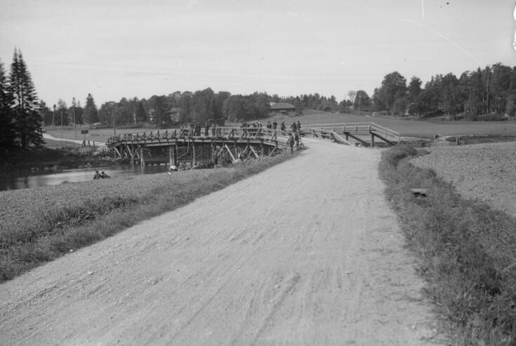 Etualalla on hiekkatie, kuvan keskellä on puusilta, jolle on kerääntynyt runsaasti väkeä katsomaan veteen. Auto oli suistunut sillan puukaiteen läpi Vantaanjokeen. 