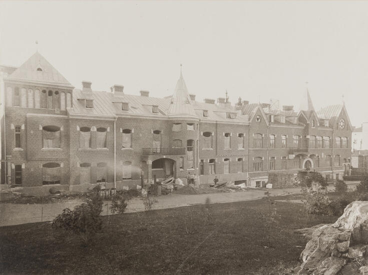 Kaksiosainen kolmikerroksinen tiilirakennus, jonka oikeanpuoleinen osa on valmis mutta vasemmanpuoleisessa ikkunoiden tilalla on säkkikangasta. Pihalla on rakennustarvikkeita.