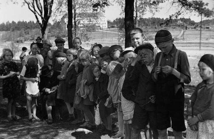 Etualalla on ryhmä lapsia kesävaatteissa. Taustalla on puita, nurmikkoa ja nelikerroksinen kivirakennus, jossa on aumakatto.