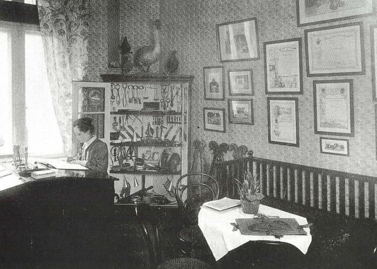 Keskellä on avoin nurkkakaappi täynnä instrumentteja. Vasemmalla istuu nainen kirjoituspöydän ääressä ikkunan luona. Oikealla on sohva.