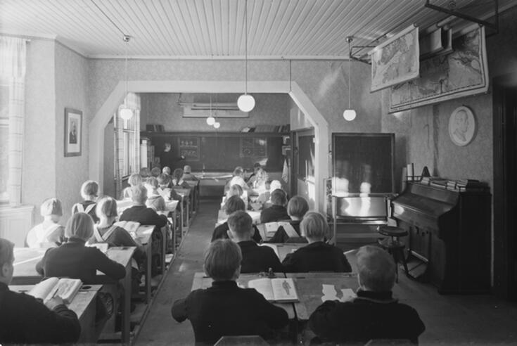 Vy framåt från den bakre delen av ett klassrum. På bilden syns rader av pulpeter där elever sitter med ryggen mot fotografen. En lärare står framför klassen och skriver på tavlan. Från taket hänger klotformade lampor. Till höger, mot väggen, står en tramporgel. Ovanför hänger kartrullar från taket.