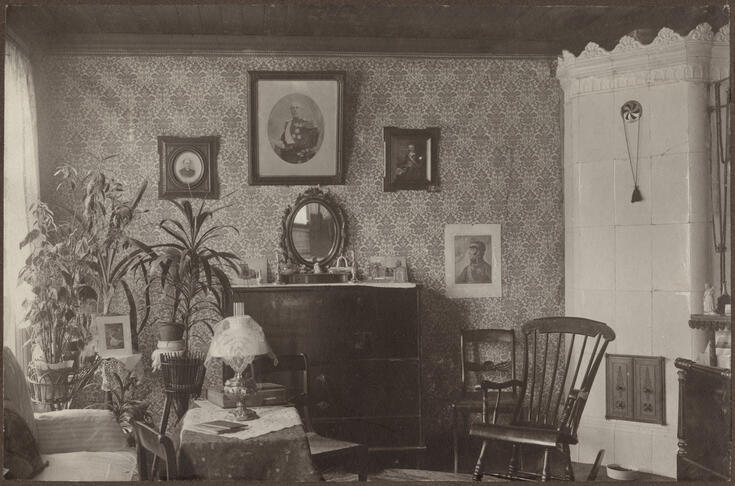 Huoneessa on kaakeliuuni, keinutuoli, kasveja sekä tauluja seinillä.