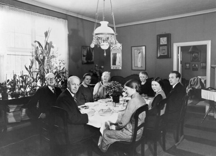 Åtta personer sitter runt ett stort matbord i mitten av rummet. Till vänster finns ett fönster som släpper in dagsljus. Det finns gröna växter framför fönstret.