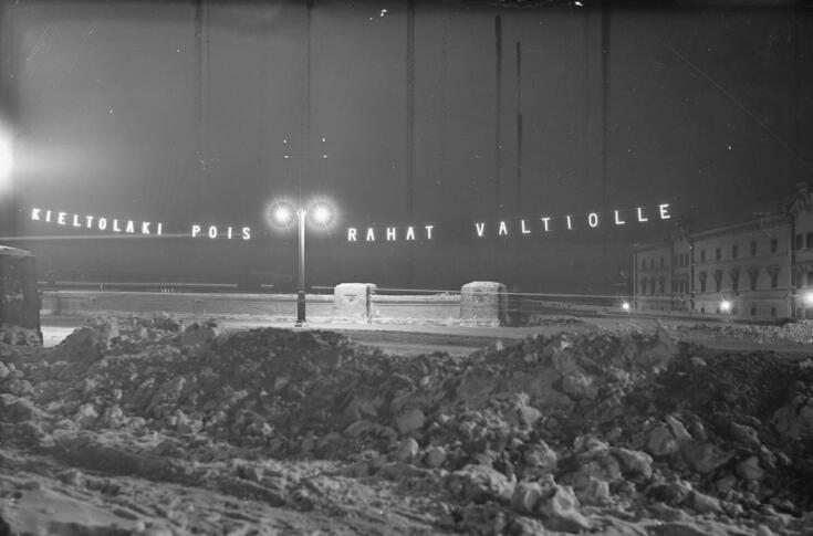 Pimeässä illassa loistavat valkoiset kirjaimet, jotka on ripustettu Pitkänsillan valopylväisiin: KIELTOLAKI POIS RAHAT VALTIOLLE