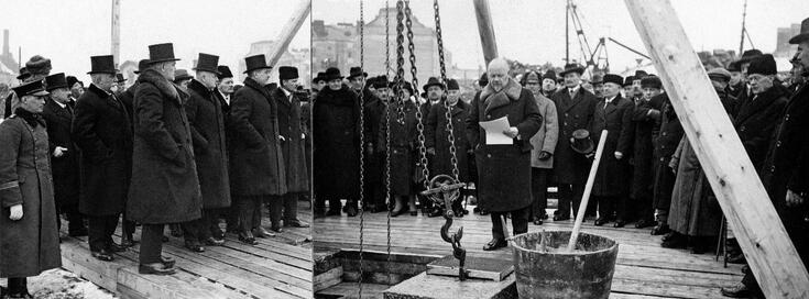 Joukko miehiä on kokoontunut tilaisuuteen, jossa lasketaan uuden eduskuntatalon kivi Arkadianmäellä. Keskellä on nosturin ketjuun kiinnitetty kivi. 