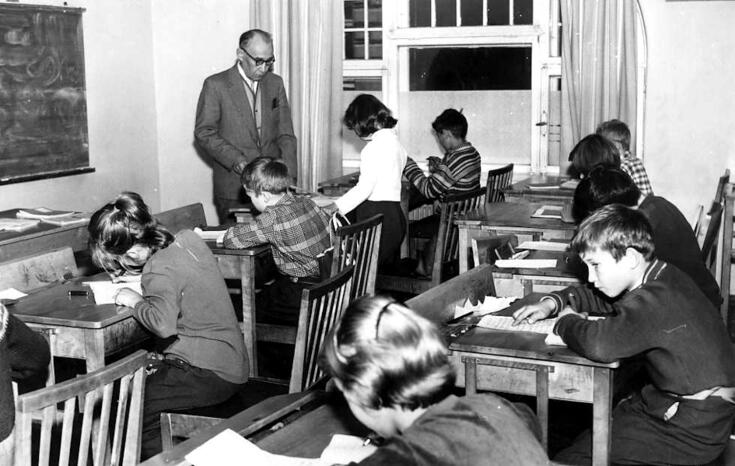 Elever i lågstadieåldern sitter vid pulpeter och arbetar. En manlig lärare står framför klassen.