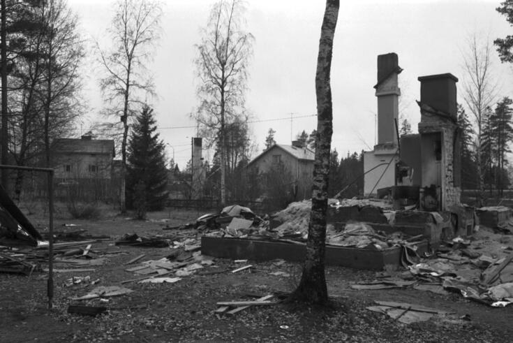 Etualalla on purettujen puutalojen jäänteitä ja niiden keskellä savuhormeja. Taustalla näkyy muutama puolitoistakerroksinen puutalo sekä koivuja. 