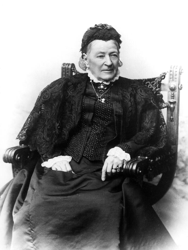 Tummiin vaatteisiin ja pitsimyssyyn pukeutunut ryhdikäs vanha nainen istuu nojatuolissa.