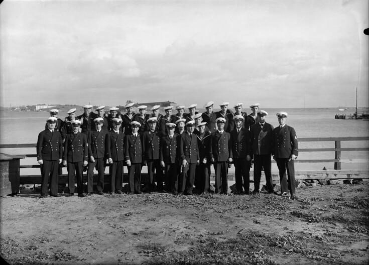 Sotilaita rivissä meren rannalla. Päällä tummat laivaston takit ja housut, päässä valkoinen laivaston lakki.