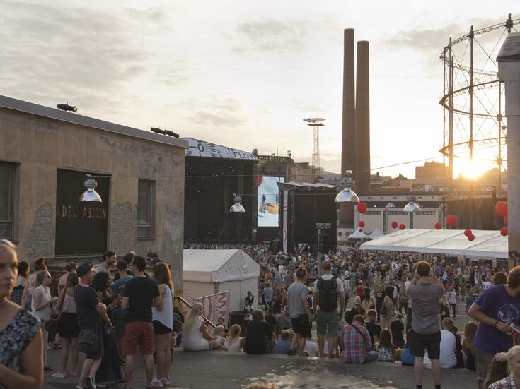 Publik på Flowfestivalen en sommarkväll i Södervik. I bakgrunden syns kraftverkets skorstenar och solen som går ner bakom gasklockans ramverk av stål.