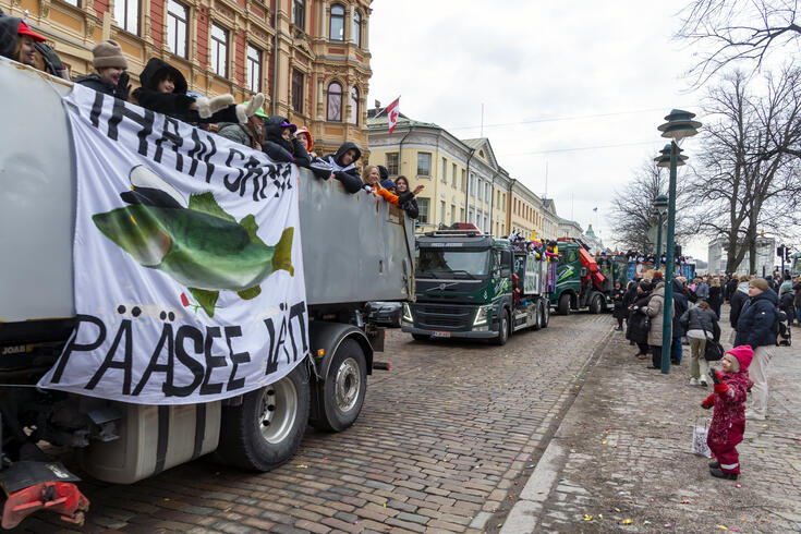 Abiturientit matkustavat penkkariasuihin pukeutuneina kuorma-autojen lavoilla. Ihmisiä on kerääntynyt seuraamaan penkkariajelua.