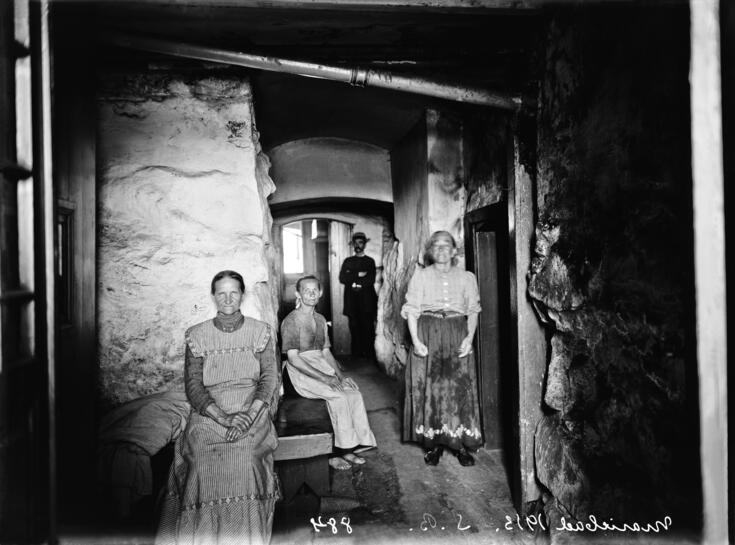 A basement room with two women sitting and one standing in the foreground and a man wearing a hat in the background.  The standing woman has wet spots on her apron.